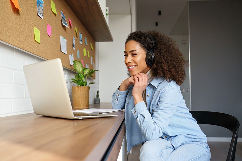 Person wearing headphones using a laptop at a desk with a corkboard and sticky notes in the background